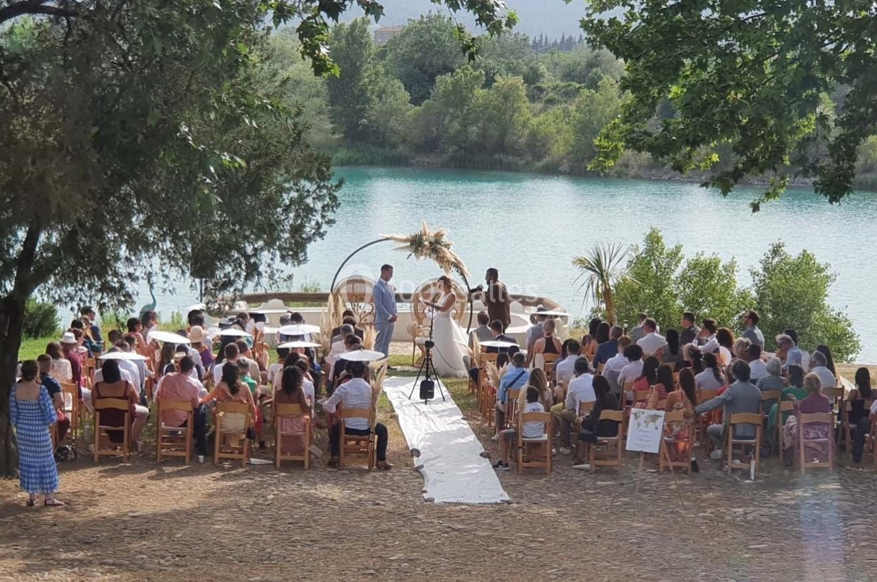 Cérémonie en plein air au bord d'un lac, avec des invités assis sous des arbres et un couple devant un autel décoré.