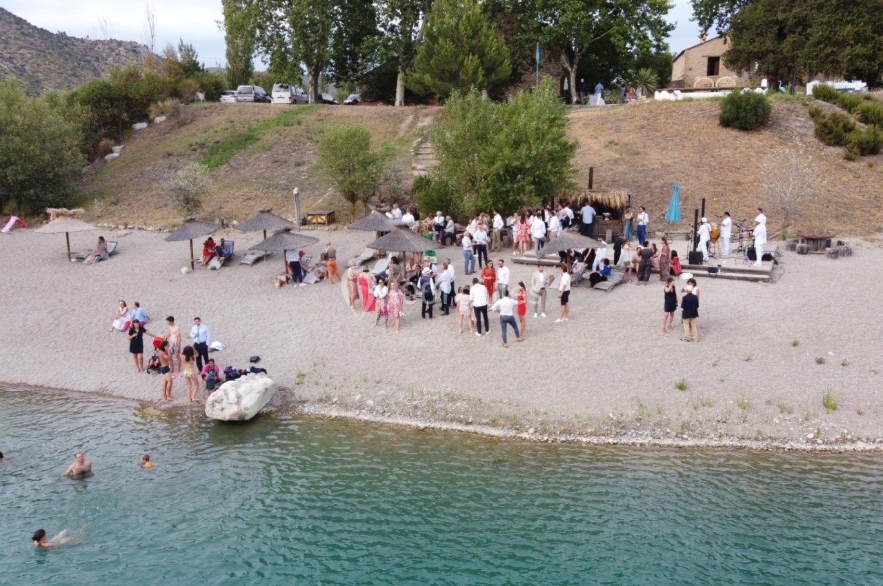 Groupe de personnes rassemblées sur une plage près d'un lac, avec des parasols et des arbres en arrière-plan.