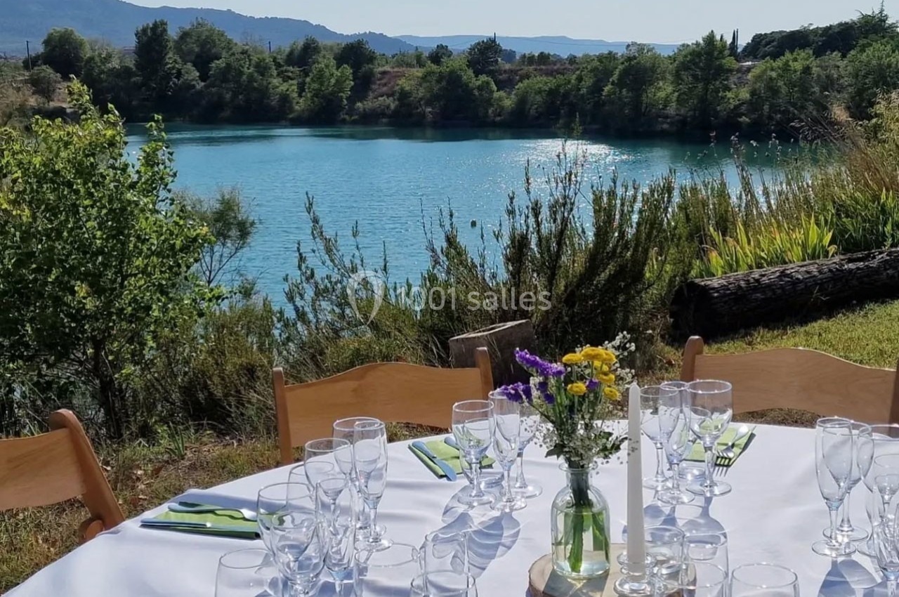 Table dressée avec nappes blanches, verres et fleurs, située en extérieur près d'un lac entouré de végétation.