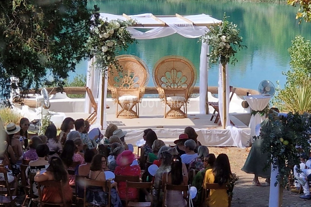 Cérémonie en plein air avec arche décorée, deux fauteuils en osier et invités assis devant un lac.