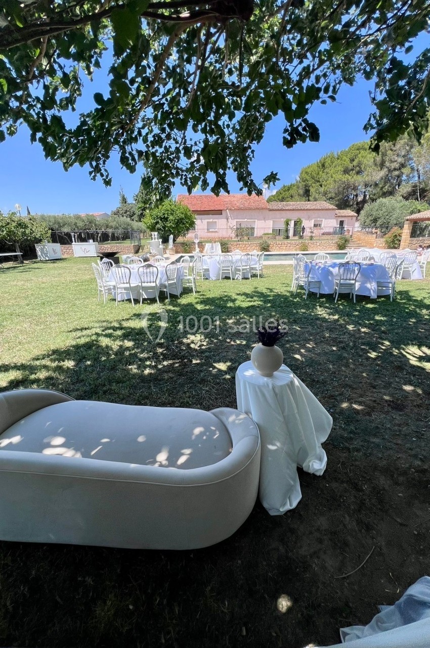 Mobilier de réception installé dans un jardin ombragé, avec des tables dressées et un canapé blanc au premier plan.