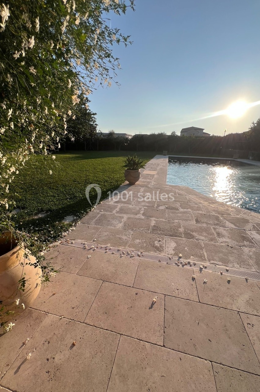 Terrasse en pierre bordant une piscine, avec pelouse, arbustes fleuris et soleil couchant à l'horizon.