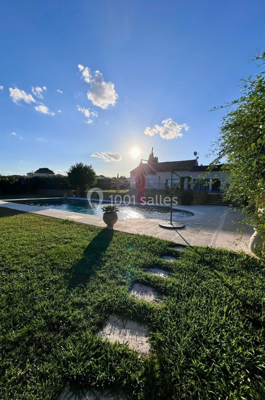 Jardin avec piscine, pelouse verte et maison en arrière-plan sous un ciel bleu ensoleillé.