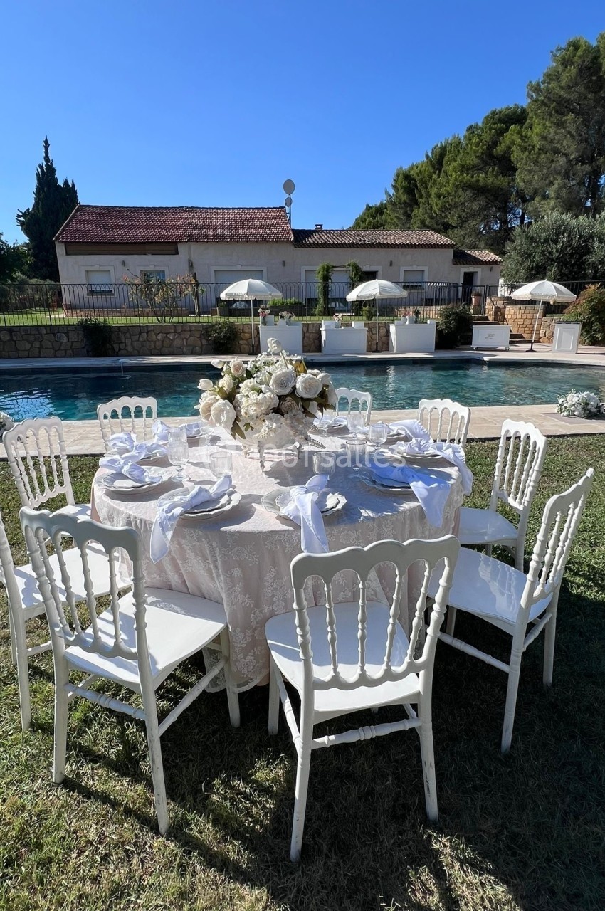 Table ronde élégamment dressée avec des chaises blanches, située dans un jardin près d'une piscine et de parasols.