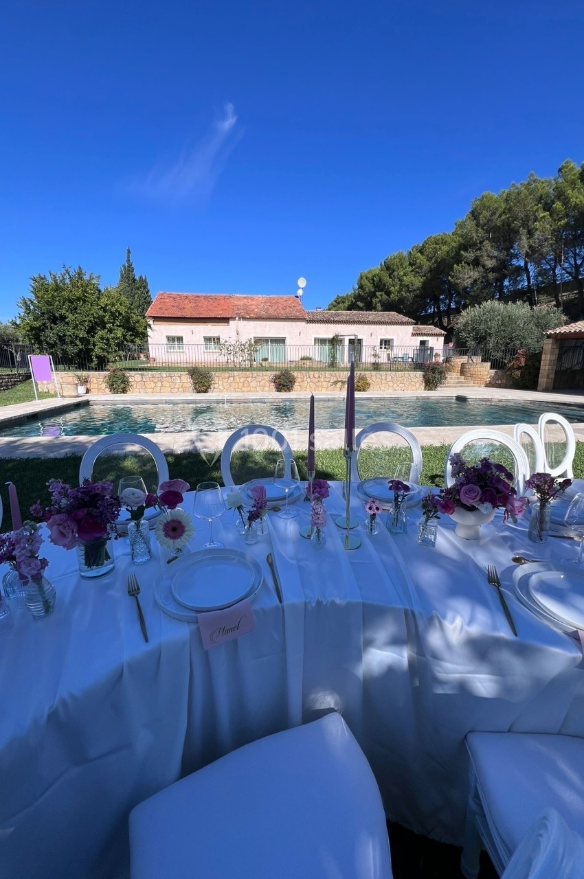 Table dressée avec nappes blanches, décorations florales et bougies, devant une piscine et un bâtiment en arrière-plan.