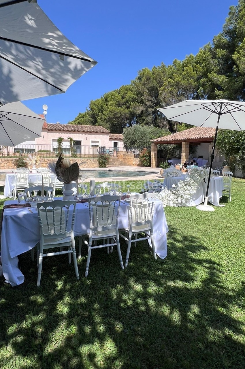 Tables décorées en blanc avec chaises disposées sur une pelouse, entourées de parasols et d'arbres sous un ciel bleu.