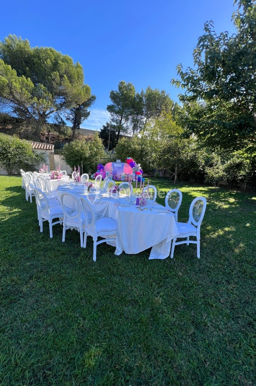 Tables blanches décorées avec des fleurs roses et des bougies, installées dans un jardin verdoyant sous un ciel bleu.