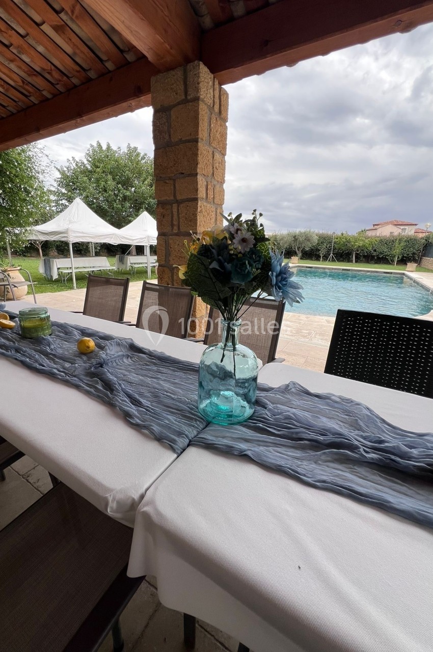 Table extérieure dressée avec une nappe blanche, un chemin de table bleu, un vase fleuri et vue sur une piscine.