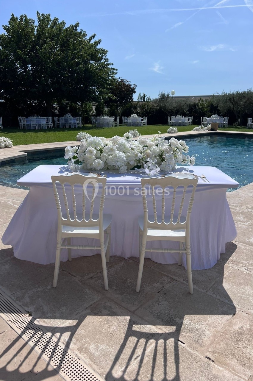 Deux chaises blanches devant une table décorée de fleurs blanches, près d'une piscine dans un jardin ensoleillé.