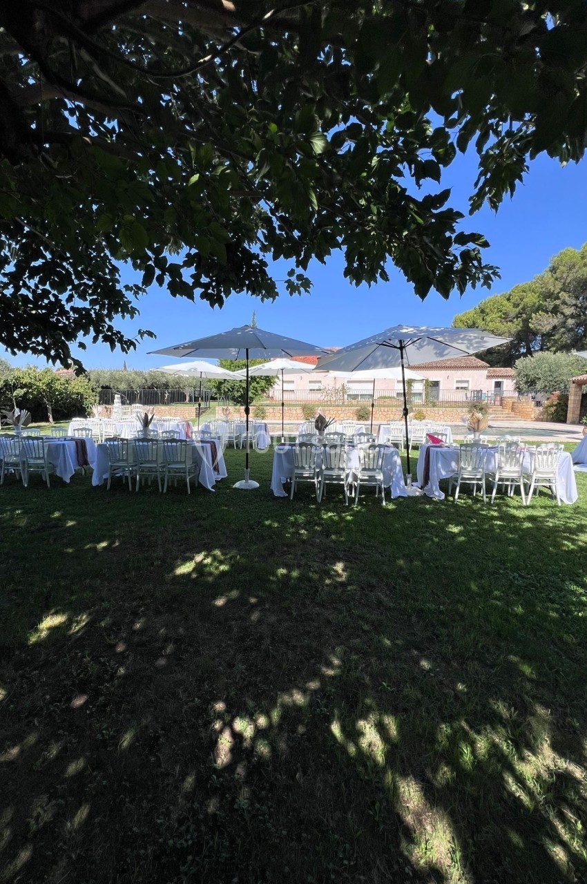 Tables et chaises blanches disposées sous des parasols dans un jardin ombragé, prêtes pour un événement en plein air.