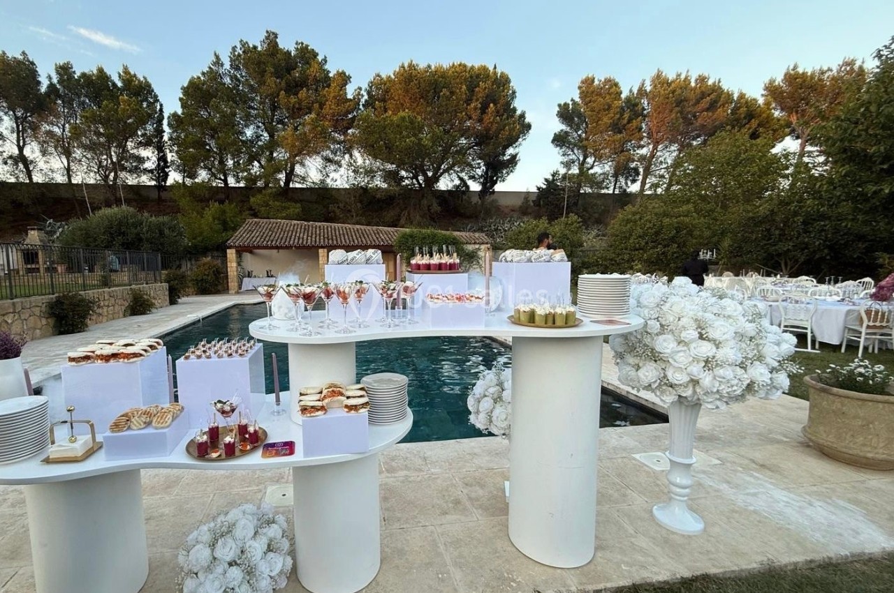 Buffet de desserts dressé en extérieur près d'une piscine, entouré de tables et de végétation.