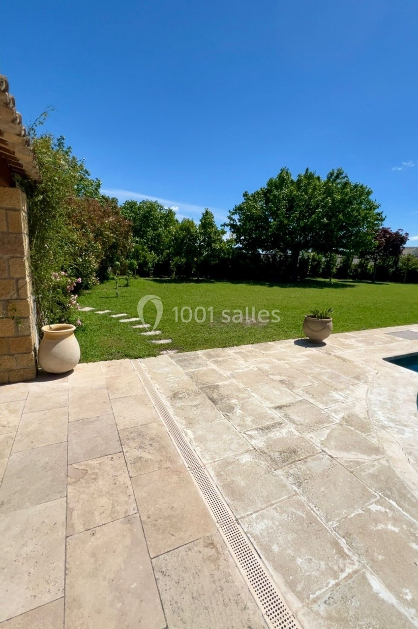 Terrasse en pierre donnant sur un jardin verdoyant avec arbres, pelouse et pots de fleurs, sous un ciel bleu dégagé.