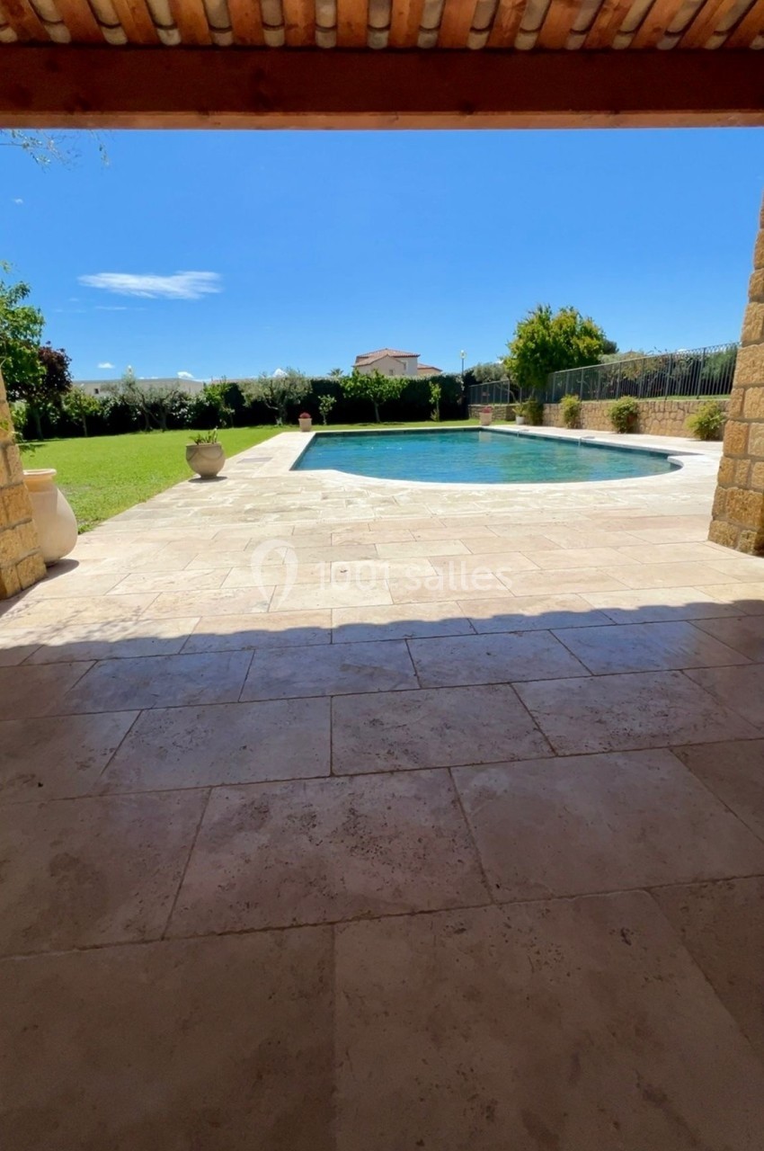 Vue depuis une terrasse couverte sur une piscine entourée de pelouse et d'arbustes sous un ciel bleu.