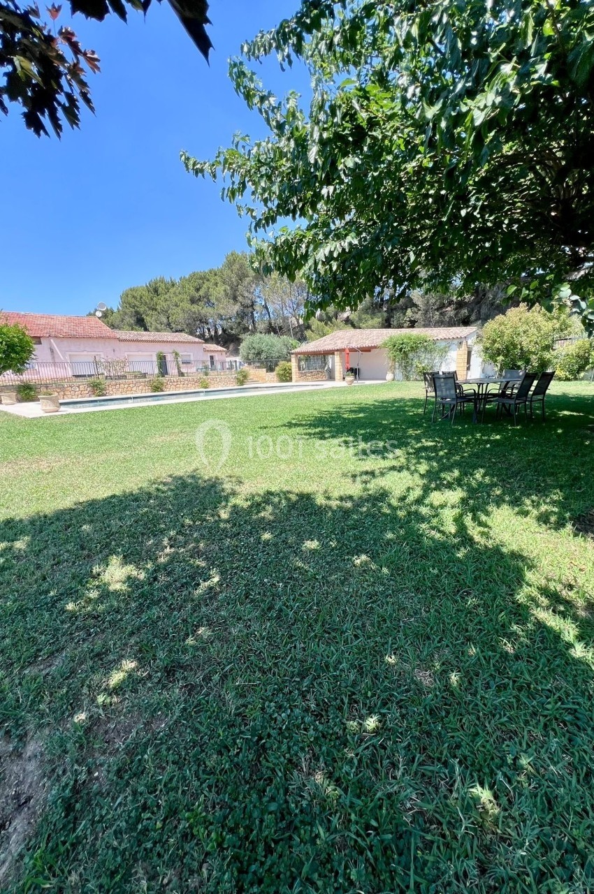 Jardin verdoyant avec pelouse, piscine, chaises sous un arbre et bâtiments en arrière-plan par une journée ensoleillée.
