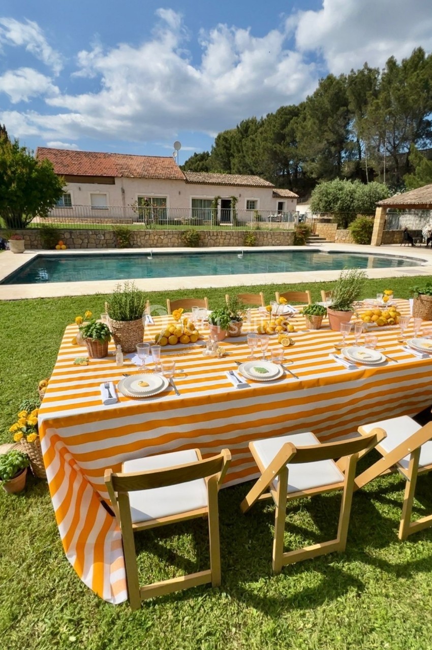 Table dressée avec une nappe rayée orange et blanche, entourée de chaises, face à une piscine dans un jardin ensoleillé.