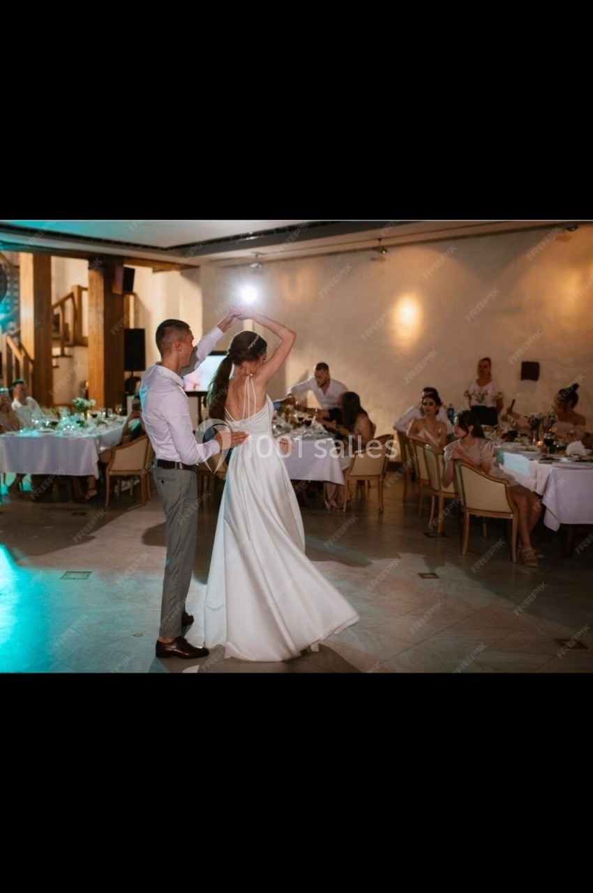 Un couple danse lors d'une réception dans une salle éclairée, entouré de tables dressées et d'invités assis.