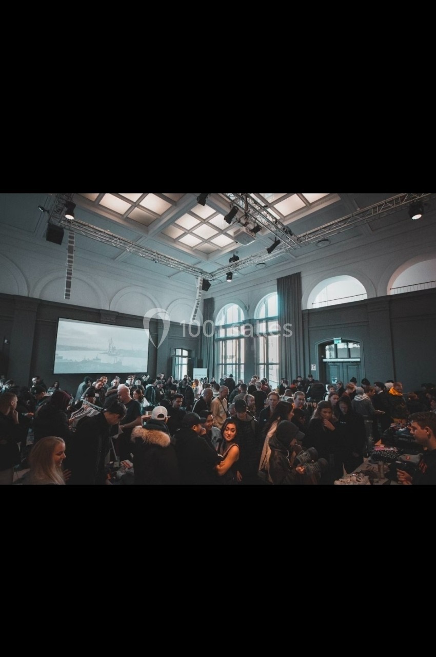 Salle bondée avec des personnes discutant et se déplaçant sous un plafond lumineux et de grandes fenêtres.