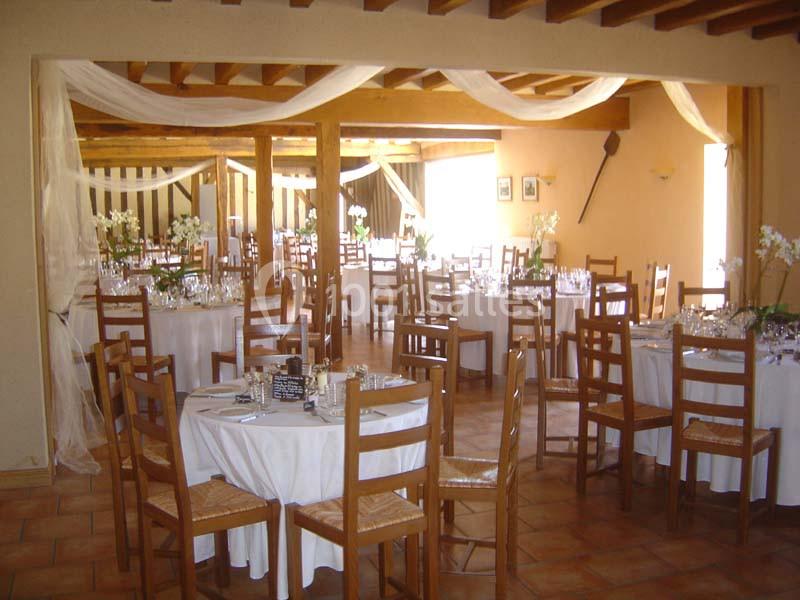 Salle de réception décorée avec des tables rondes dressées, nappes blanches et chaises en bois, ornée de voilages.