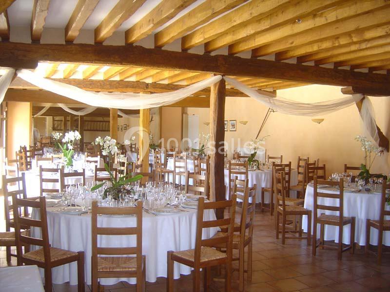 Salle de réception décorée avec des tables rondes, nappes blanches, chaises en bois et arrangements floraux.