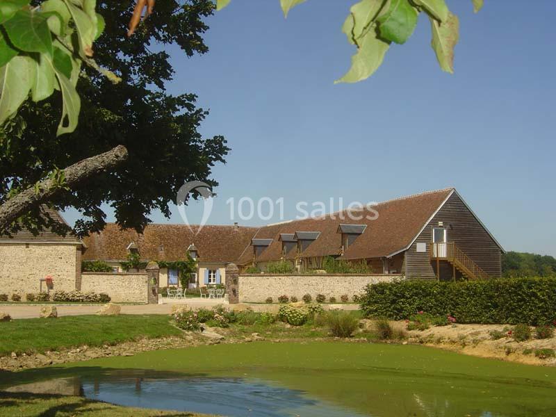 Bâtiment rural en pierre et bois avec toit en pente, entouré de verdure et d'un petit étang au premier plan.