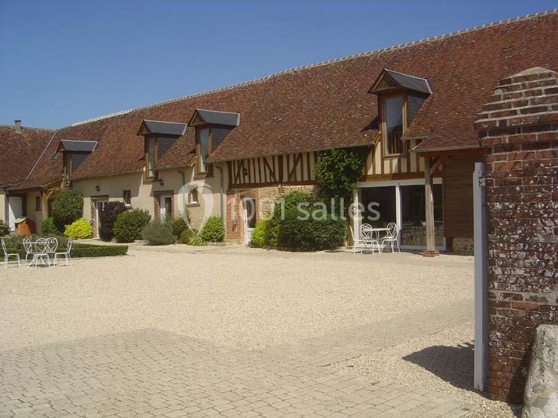 Grande bâtisse en pierre et colombages avec toit en tuiles, entourée d'une cour pavée et de végétation.
