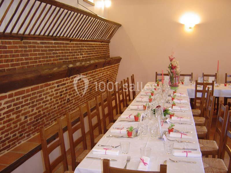 Salle avec une longue table dressée pour un repas, entourée de chaises en bois, et décorée de fleurs et serviettes pliées.