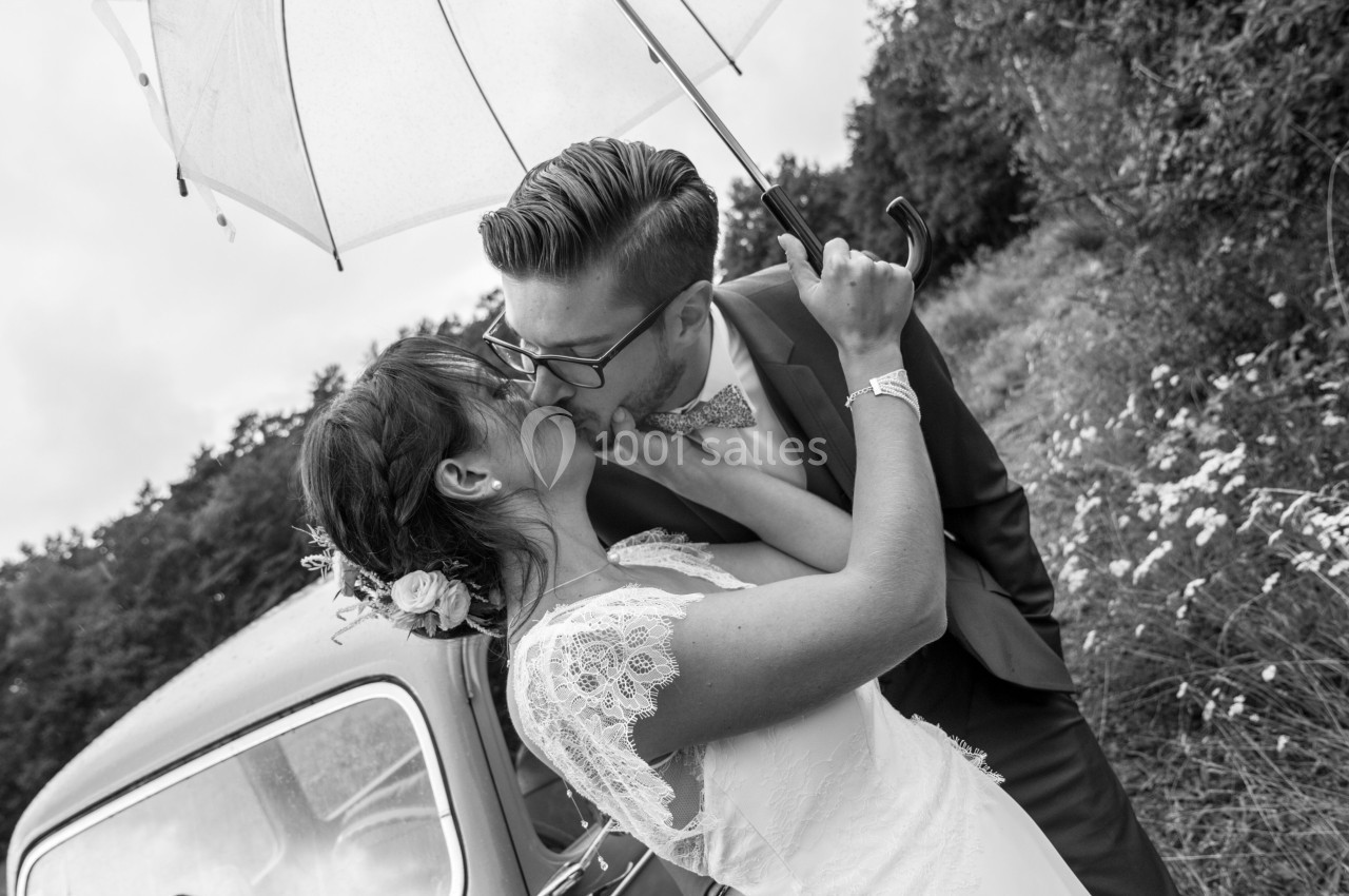 Un couple en tenue de mariage s'embrasse sous un parapluie transparent près d'une voiture ancienne dans un cadre champêtre.