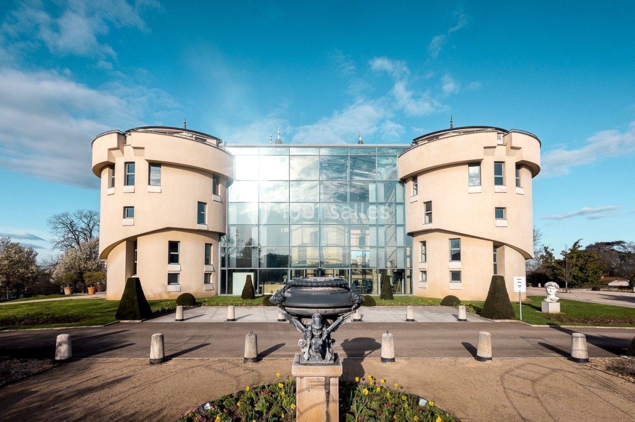 Façade d'un bâtiment moderne en verre entouré de deux tours rondes, avec une allée et des fleurs au premier plan.