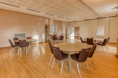 Salle de restaurant lumineuse avec de grandes baies vitrées, des tables dressées et un olivier en pot au centre.