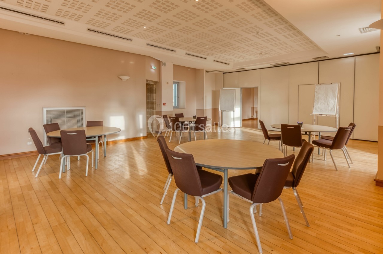 Salle lumineuse avec parquet, tables rondes entourées de chaises marron et panneaux amovibles en arrière-plan.