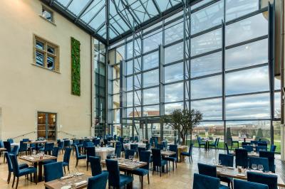 Salle de restaurant lumineuse avec de grandes baies vitrées, des tables dressées et un olivier en pot au centre.