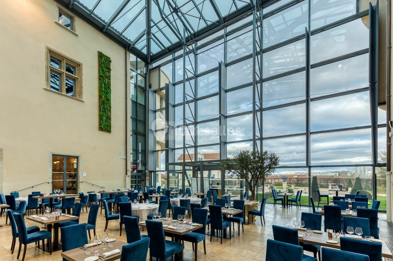 Salle de restaurant lumineuse avec de grandes baies vitrées, des tables dressées et des chaises bleues.