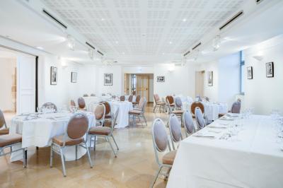 Salle de restaurant lumineuse avec de grandes baies vitrées, des tables dressées et un olivier en pot au centre.