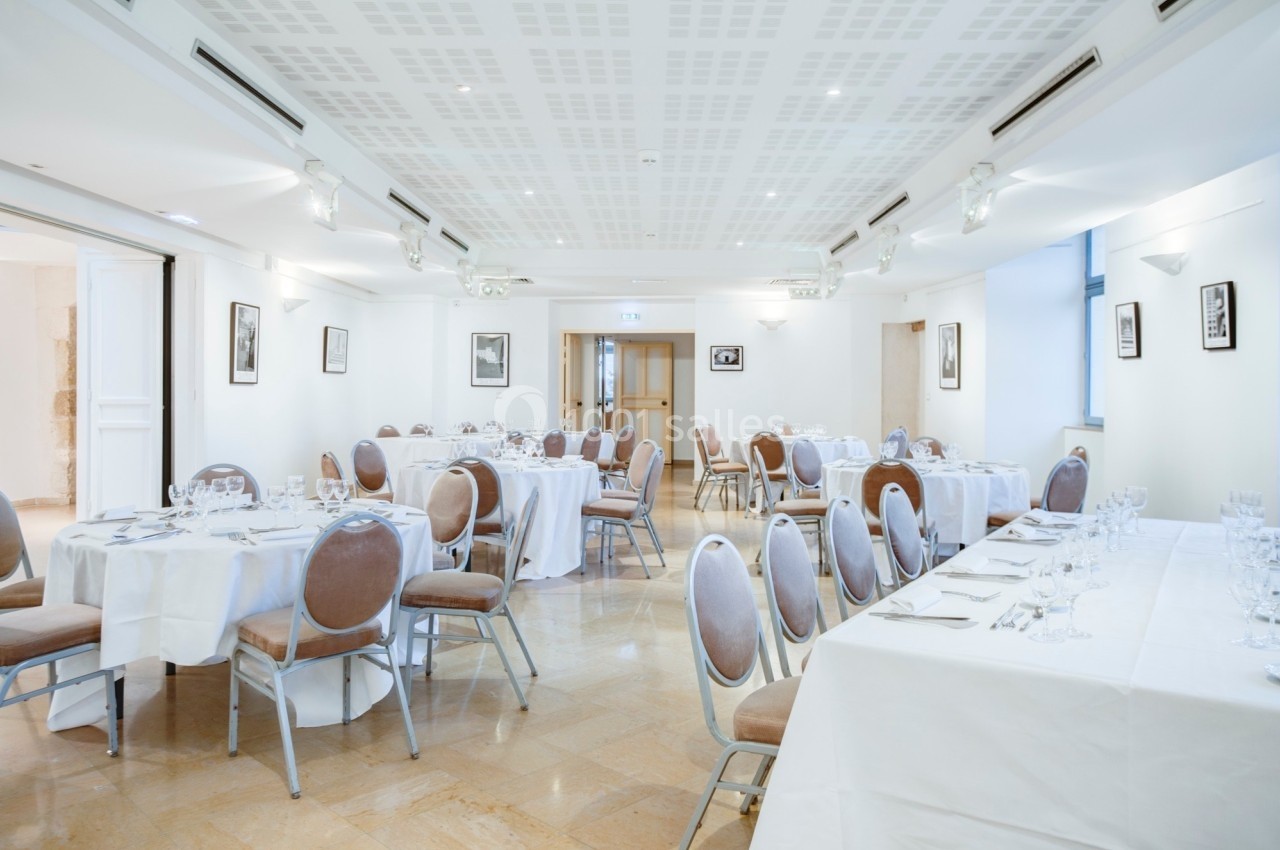 Salle lumineuse avec tables rondes dressées pour un repas, chaises rembourrées et décor minimaliste.
