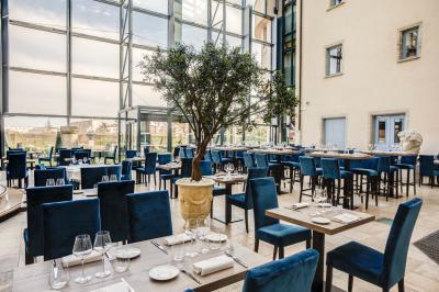 Salle de restaurant lumineuse avec de grandes baies vitrées, des tables dressées et un olivier en pot au centre.