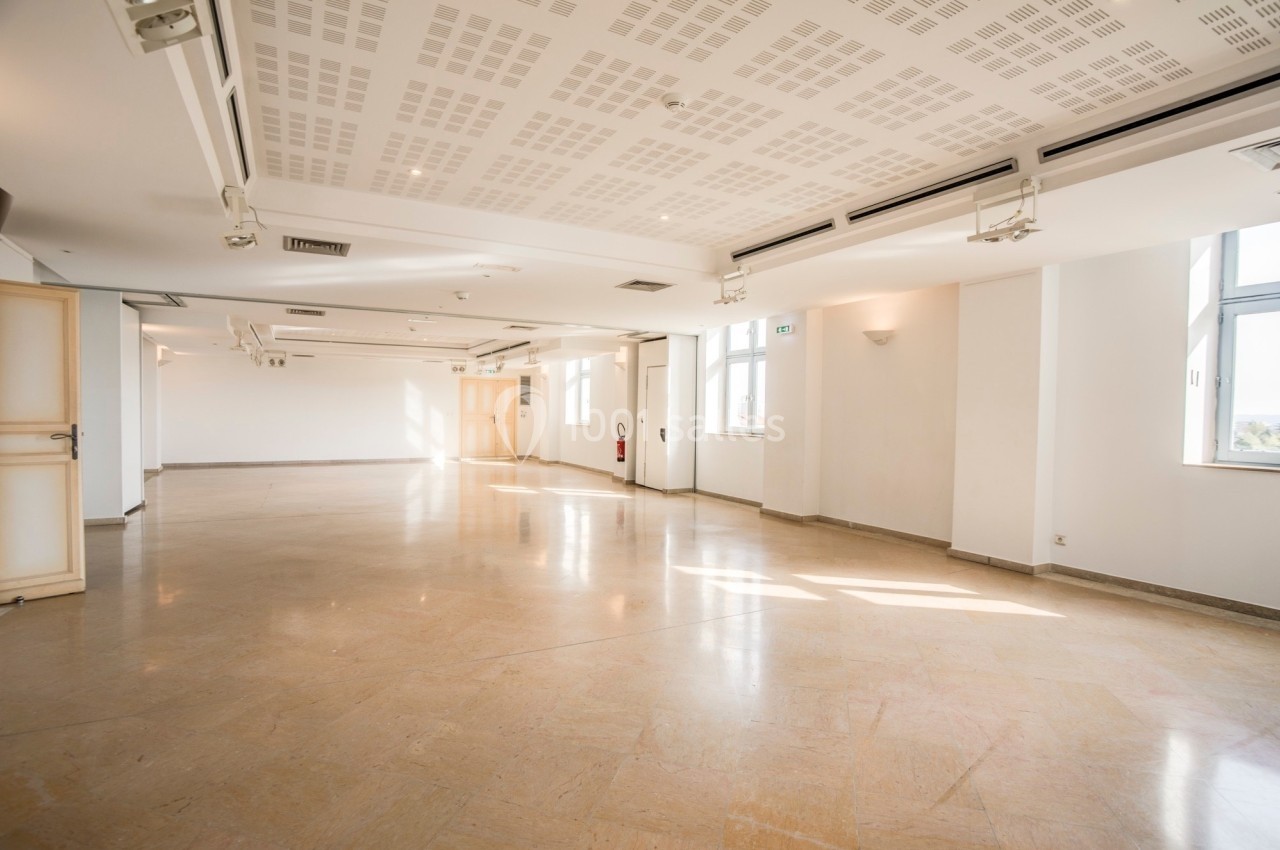 Salle lumineuse et vide avec sol en marbre, murs blancs, grandes fenêtres et plafond équipé de spots encastrés.