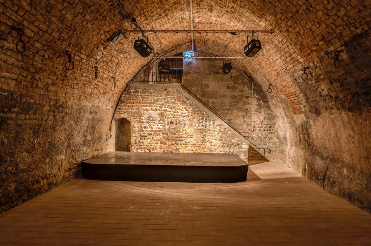 Salle voûtée en briques avec un sol carrelé, une petite scène surélevée et un éclairage discret.