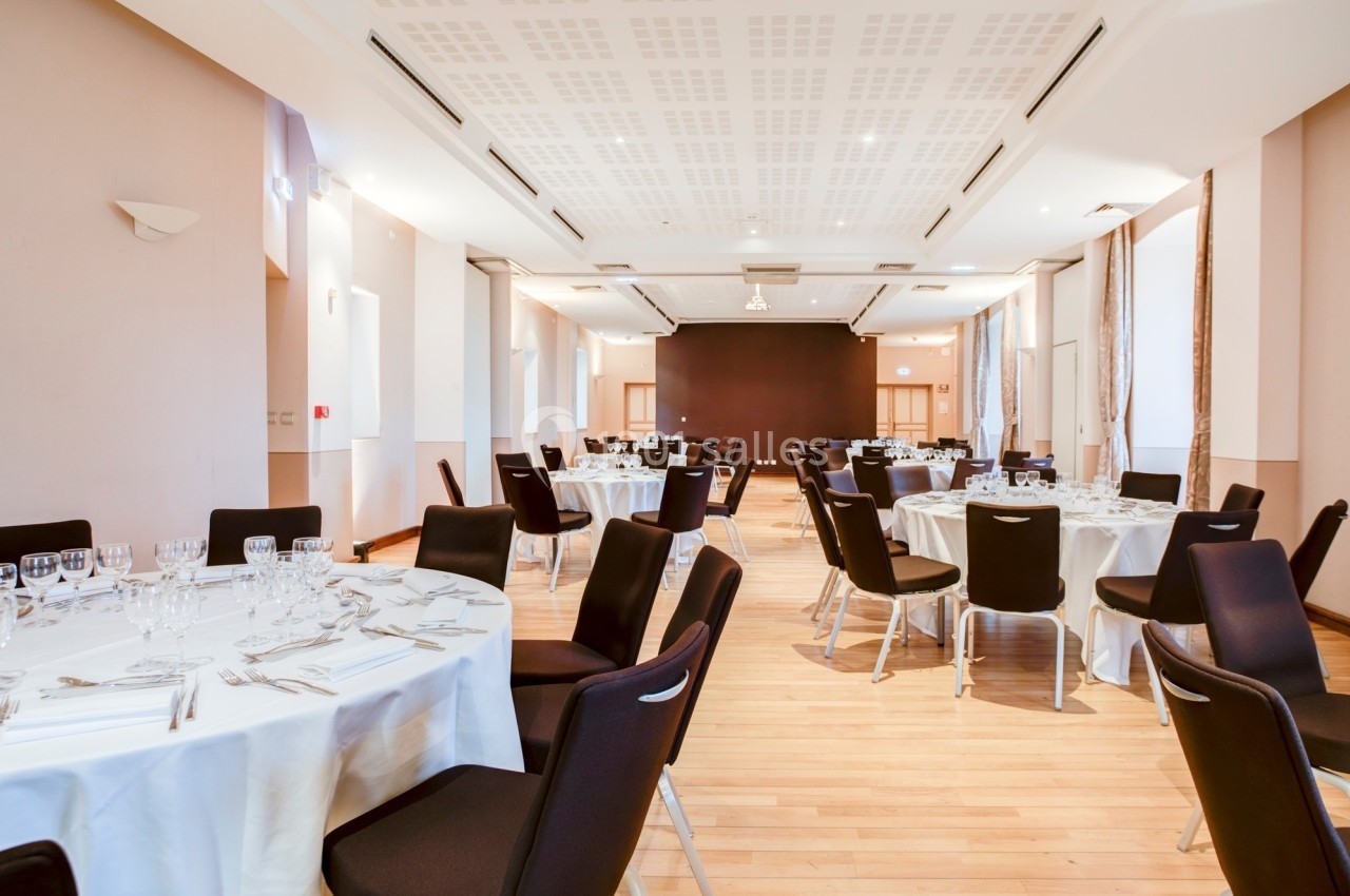 Salle de réception lumineuse avec tables rondes dressées de nappes blanches, entourées de chaises noires.