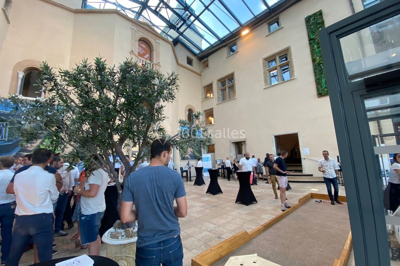 Personnes rassemblées dans une cour intérieure lumineuse avec un toit en verre, des tables hautes et un terrain de jeu.