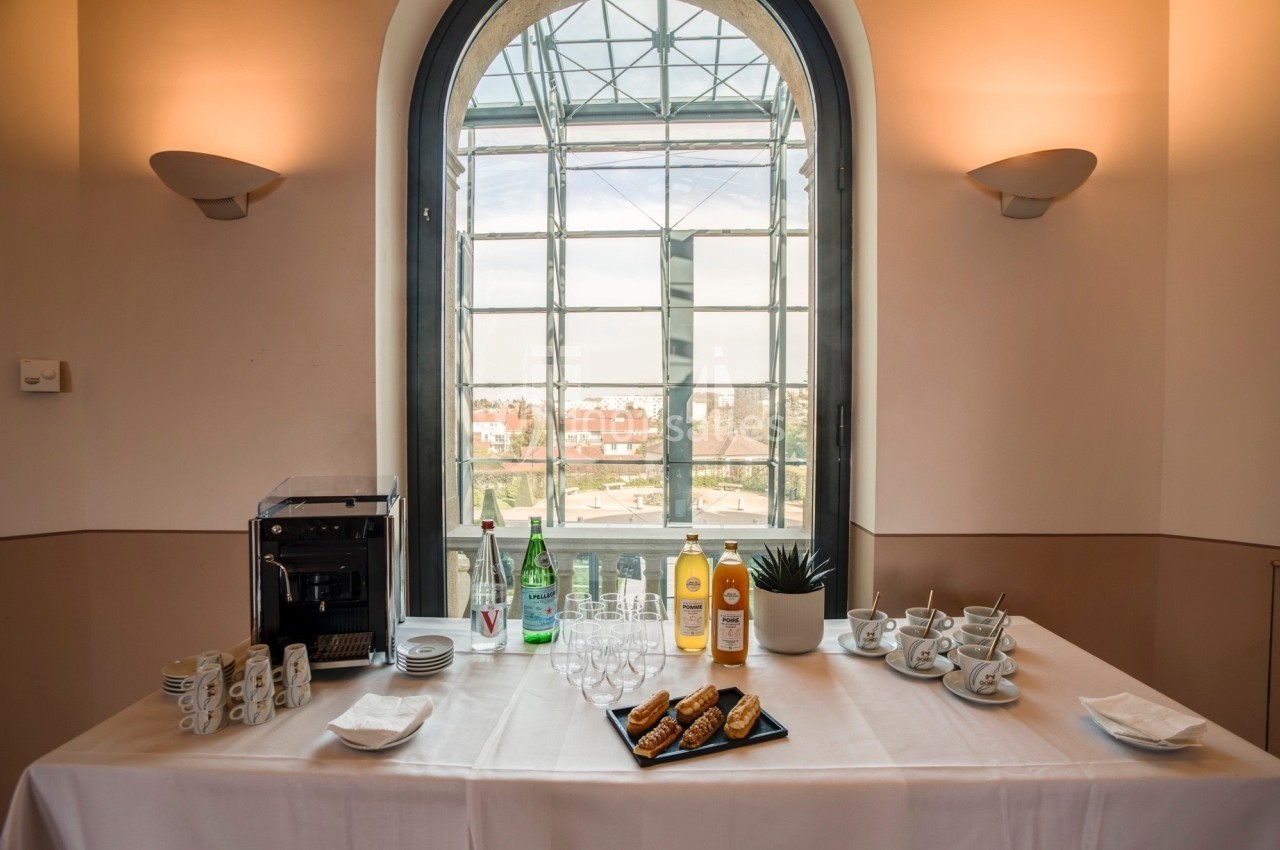 Table dressée avec machine à café, bouteilles, verres, tasses, viennoiseries et vue sur un paysage urbain par une grande…