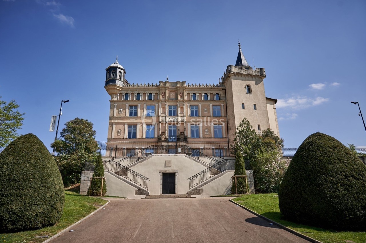 Façade d'un bâtiment historique avec tourelles, entouré de haies taillées et d'un ciel dégagé.