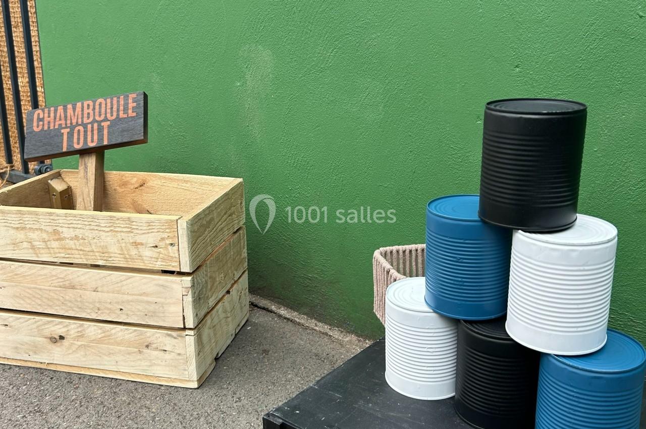 Boîte en bois avec un panneau ’Chamboule Tout’ et pyramide de boîtes de conserve colorées devant un mur vert.