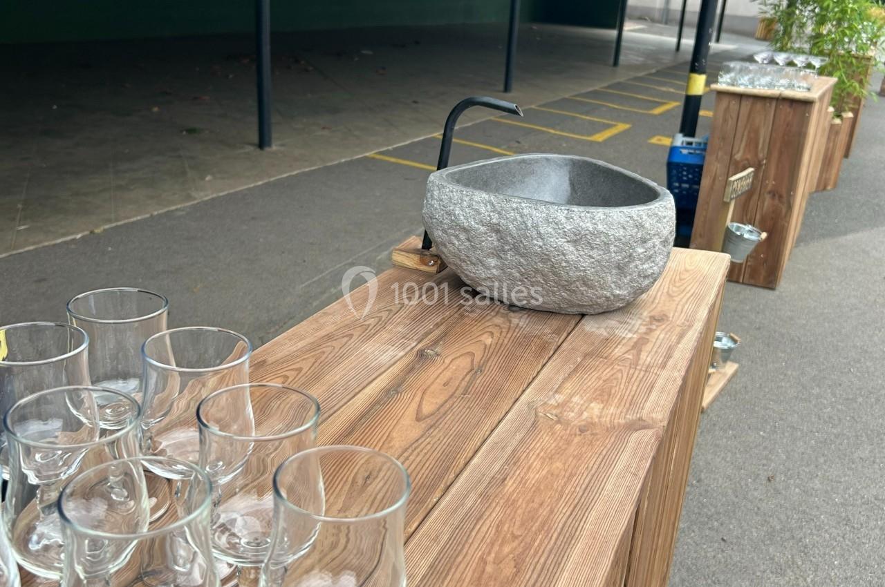Évier en pierre avec robinet noir sur un comptoir en bois, entouré de verres et situé en extérieur.