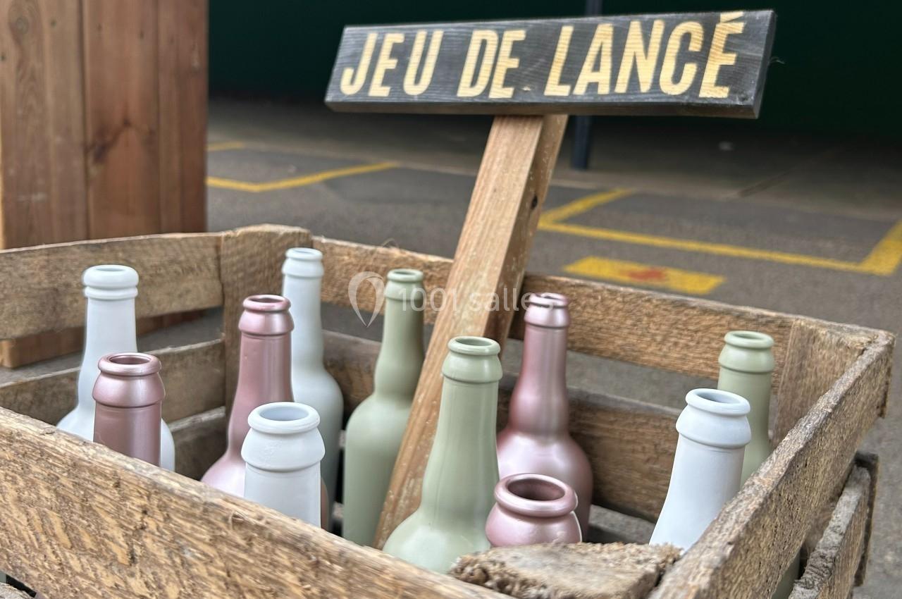Caisse en bois contenant des bouteilles colorées sous un panneau ’Jeu de lance’ dans un espace extérieur.