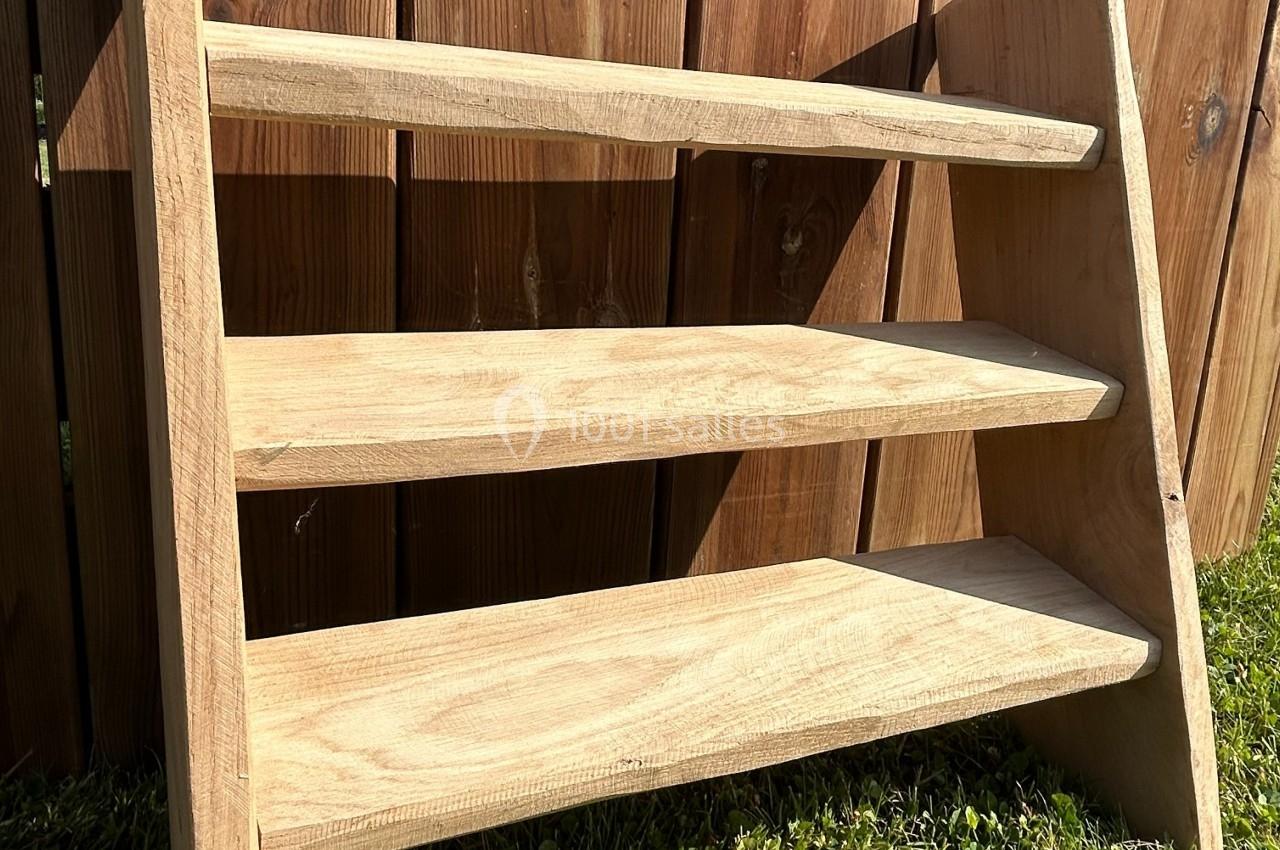 Étagère en bois brut à trois niveaux, posée devant une clôture en bois sur une pelouse.