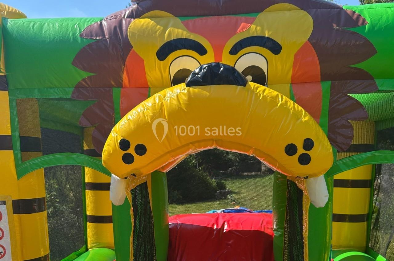 Structure gonflable colorée en forme de tête de lion avec des crocs, installée en extérieur sur une pelouse.