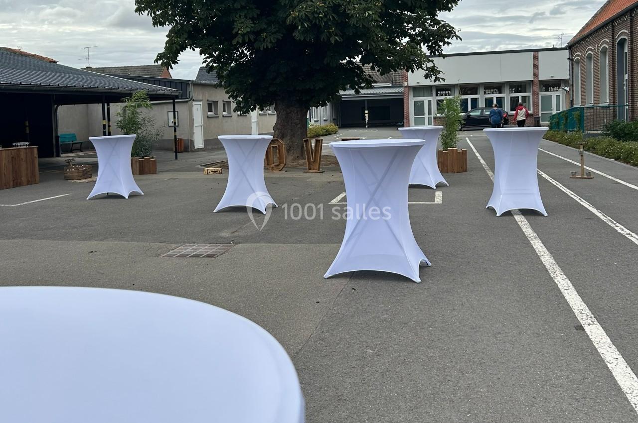 Tables hautes recouvertes de nappes blanches disposées en extérieur sur un parking devant des bâtiments.