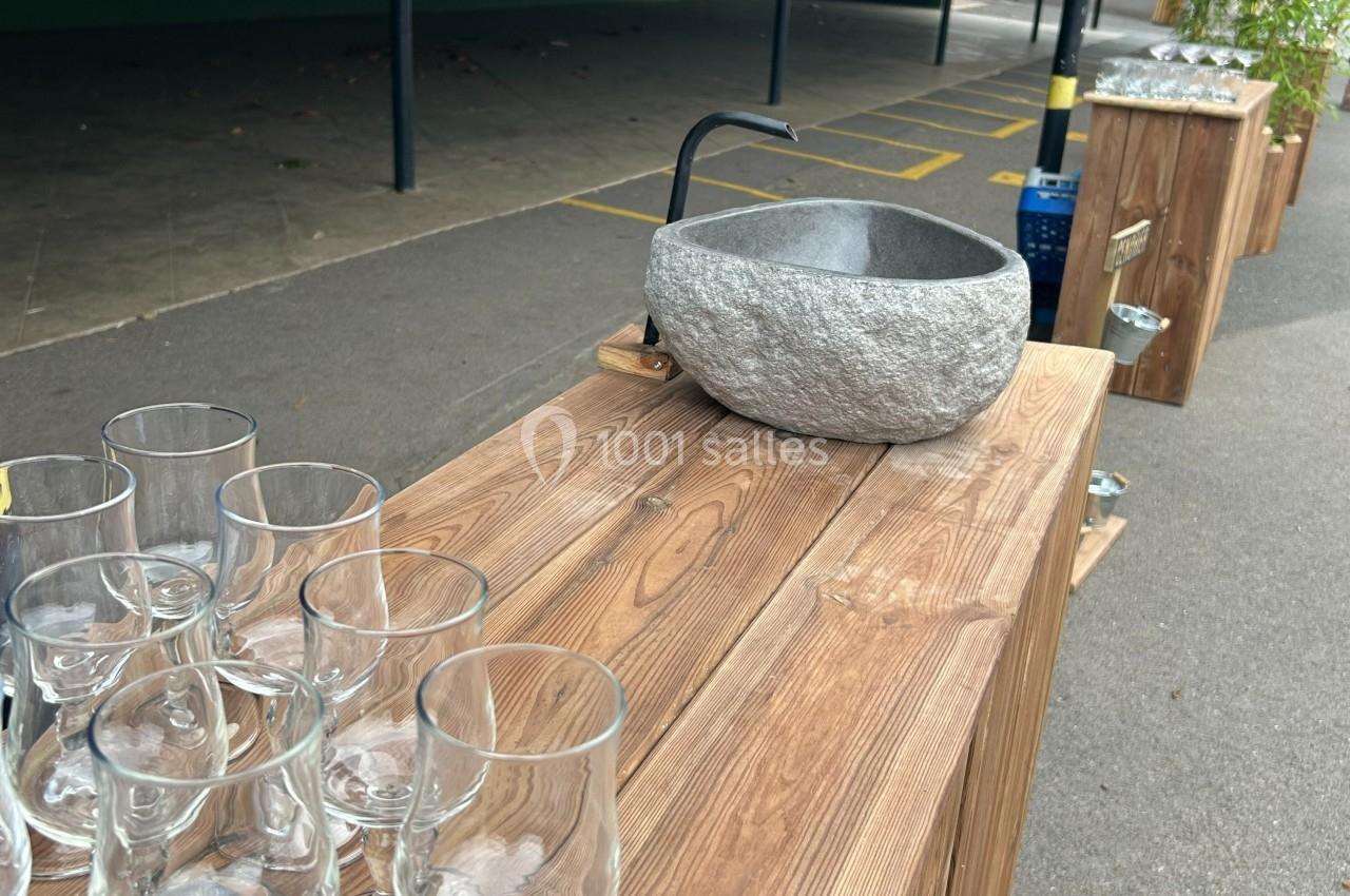 Bol en pierre avec un robinet sur une table en bois, entouré de verres transparents, en extérieur.