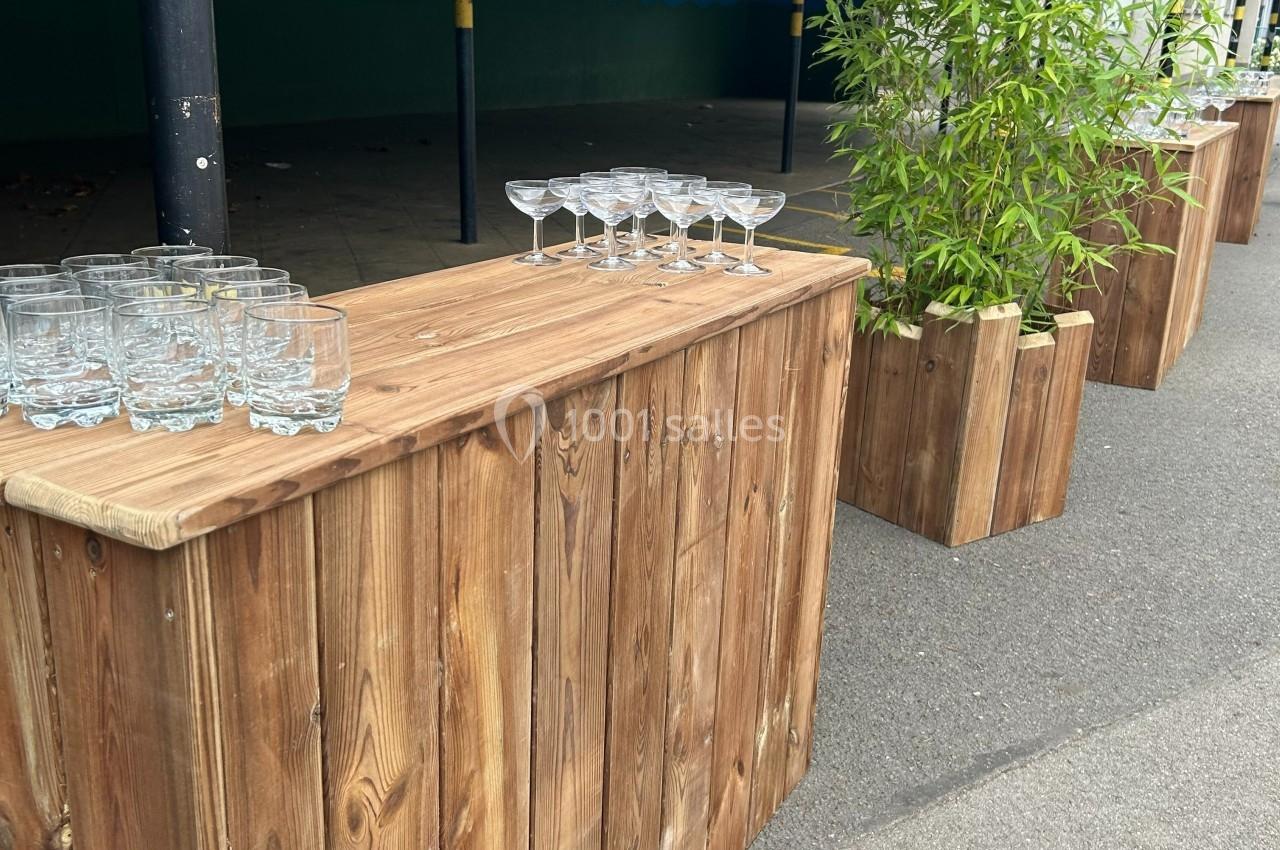 Comptoirs en bois avec des verres disposés, entourés de plantes en pot, dans un espace extérieur.