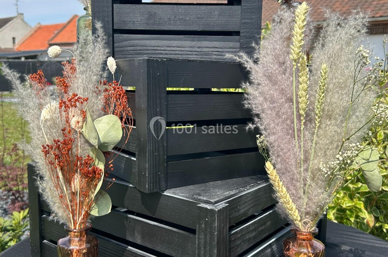 Deux caisses en bois noir empilées entourées de bouquets de fleurs séchées dans des vases en verre, en extérieur.